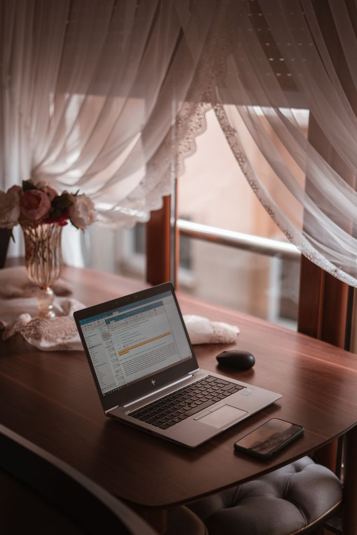 Cozy home office setup with a laptop on a wooden desk beside sheer curtains, offering a warm atmosphere.